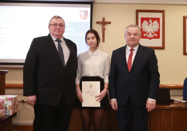 Trzy osoby stoją w sali sesyjnej urzędu powiatowego; pośrodku młoda kobieta w białej koszuli trzyma teczkę z herbem Powiatu Płockiego, a po bokach stoją dwaj mężczyźni w garniturach.