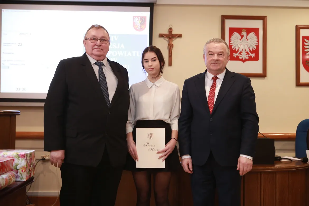 Trzy osoby stoją w sali sesyjnej urzędu powiatowego; pośrodku młoda kobieta w białej koszuli trzyma teczkę z herbem Powiatu Płockiego, a po bokach stoją dwaj mężczyźni w garniturach.