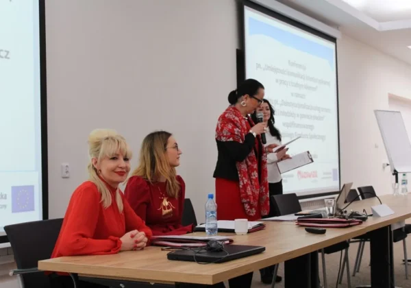 Cztery kobiety prowadzą panel konferencji na temat umiejętności komunikacji interdyscyplinarnej w pracy z trudnym klientem, siedząc i stojąc przy stole z laptopami i mikrofonami, przed projekcją slajdów w tle.