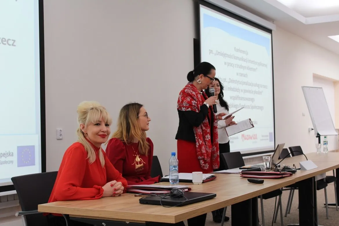 Cztery kobiety prowadzą panel konferencji na temat umiejętności komunikacji interdyscyplinarnej w pracy z trudnym klientem, siedząc i stojąc przy stole z laptopami i mikrofonami, przed projekcją slajdów w tle.