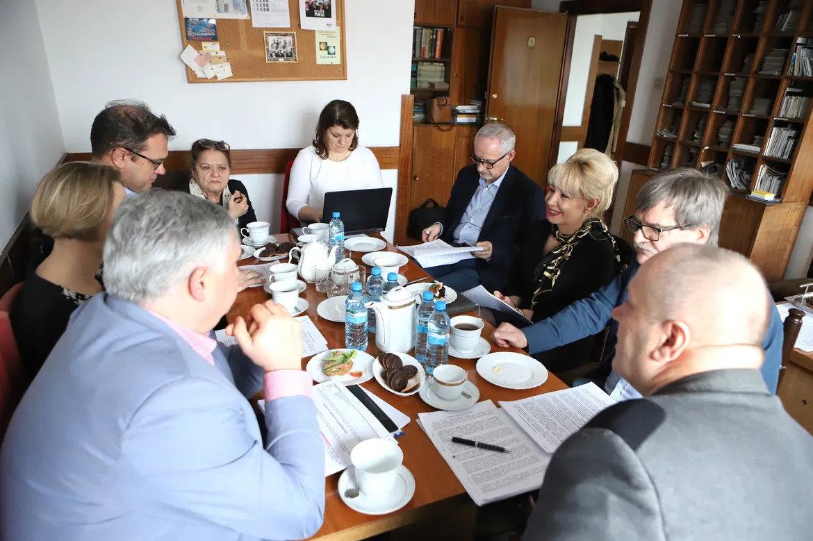 Grupa osób siedzi przy długim stole w biurze, omawiając dokumenty podczas spotkania, z napojami i przekąskami na stole, w otoczeniu regałów z książkami.