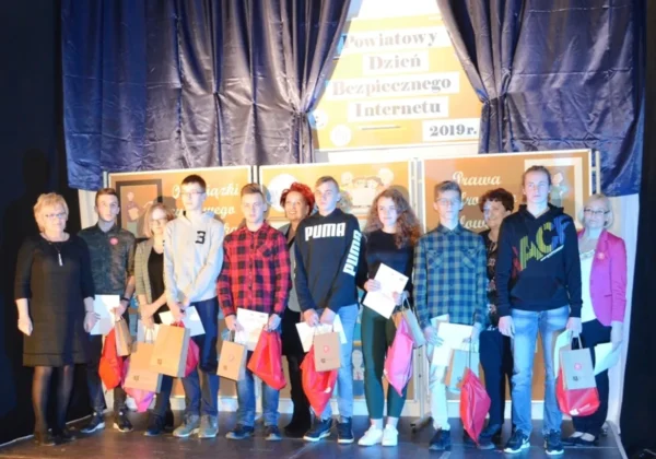 Grupa uczniów i nauczycieli stoi na scenie podczas Powiatowego Dnia Bezpiecznego Internetu w Zespole Szkół im. Stanisława Staszica w Gąbinie, trzymając torby z upominkami.