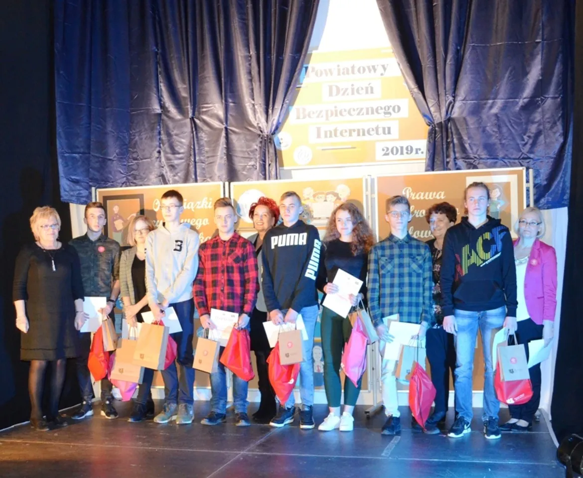 Grupa uczniów i nauczycieli stoi na scenie podczas Powiatowego Dnia Bezpiecznego Internetu w Zespole Szkół im. Stanisława Staszica w Gąbinie, trzymając torby z upominkami.