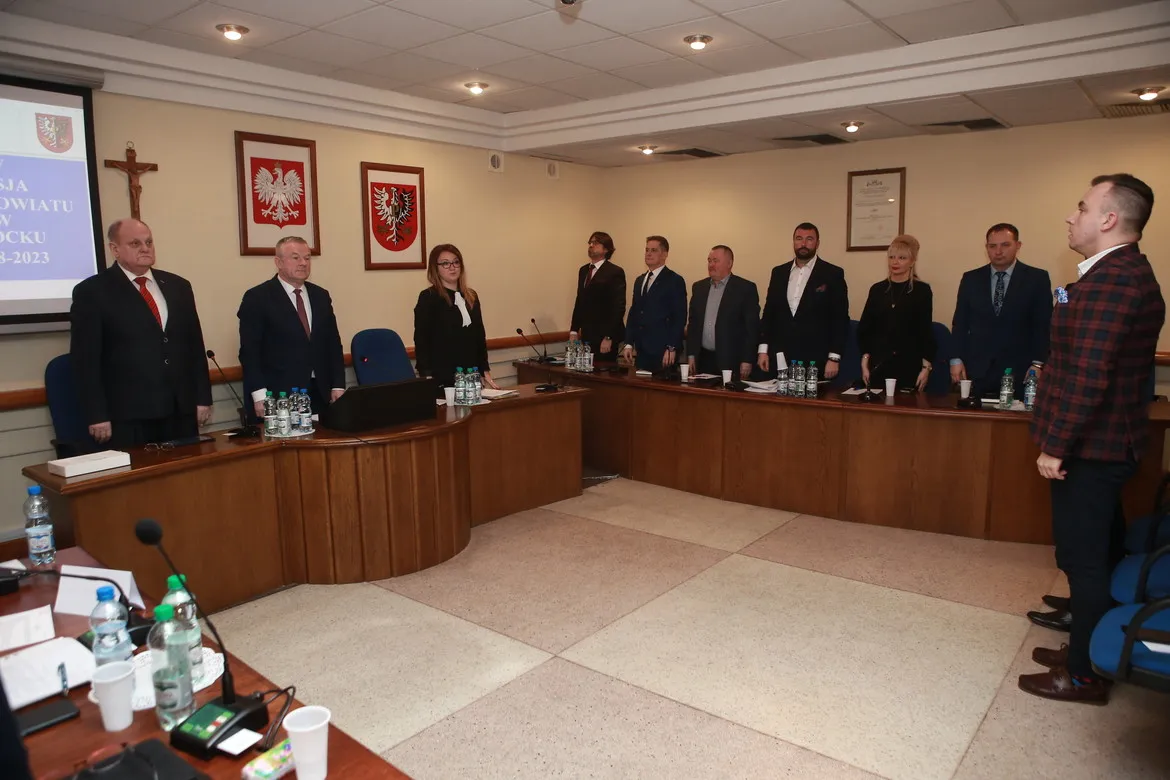 Grupa radnych stoi przy drewnianych biurkach w sali obrad podczas V sesji Rady Powiatu w Płocku.