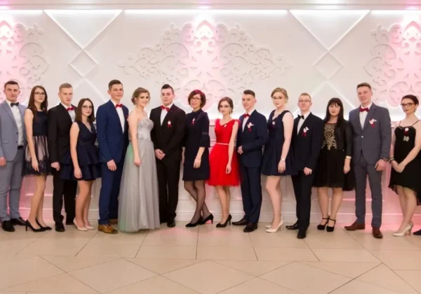 Grupa młodzieży w galowych ubraniach stoi w jednym rzędzie na tle ozdobionej ścianie podczas obchodów 100 dni do matury.