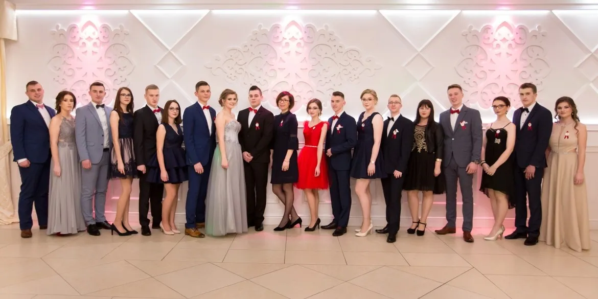 Grupa młodzieży w galowych ubraniach stoi w jednym rzędzie na tle ozdobionej ścianie podczas obchodów 100 dni do matury.