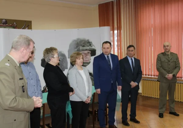 Grupa osób w mundurach i garniturach stoi w sali obok baneru z wizerunkiem żołnierza, podczas rozpoczęcia kwalifikacji wojskowej w 2019 roku.