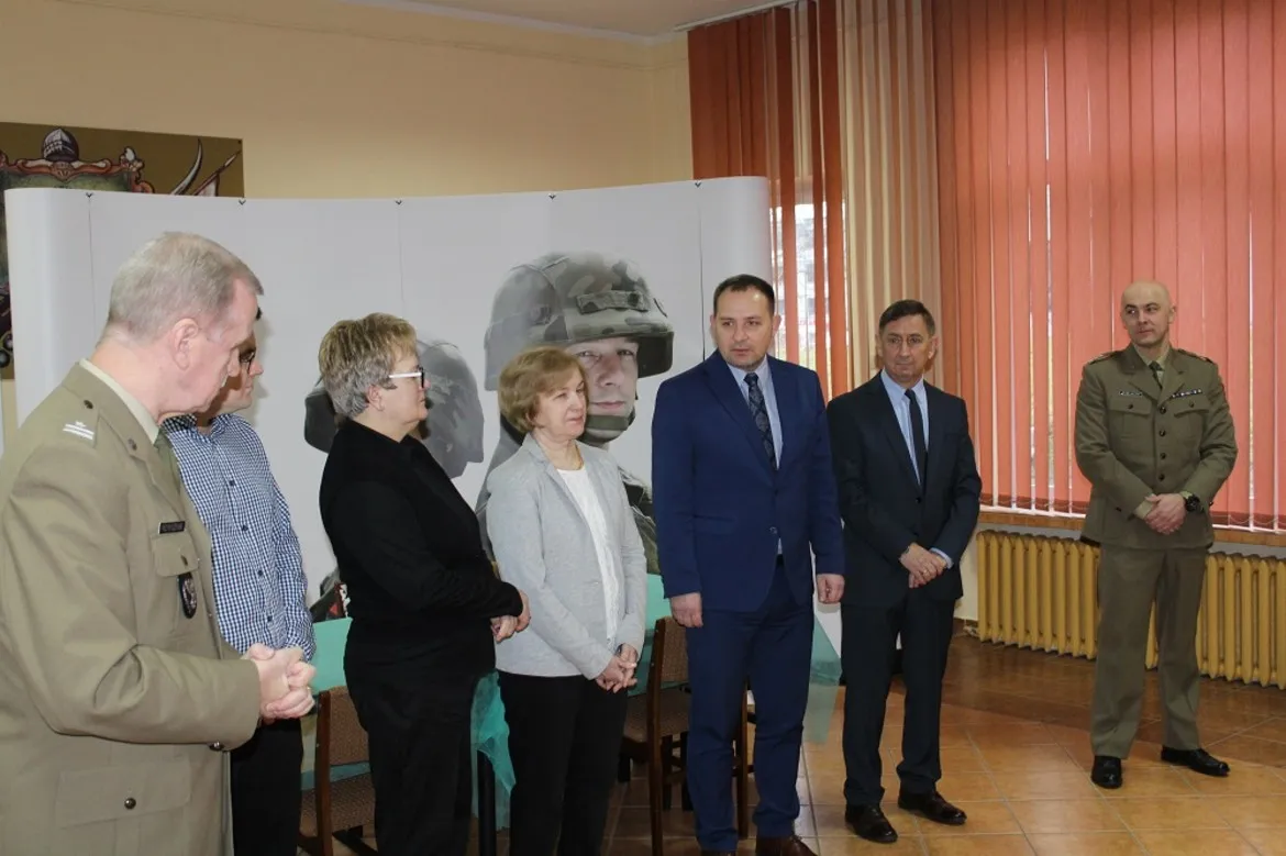 Grupa osób w mundurach i garniturach stoi w sali obok baneru z wizerunkiem żołnierza, podczas rozpoczęcia kwalifikacji wojskowej w 2019 roku.