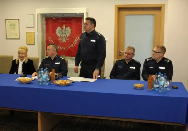 Płoccy policjanci podczas konferencji w sali urzędu omawiają stan bezpieczeństwa w minionym roku, siedząc przy stole z wodą i przekąskami, z flagą w tle.