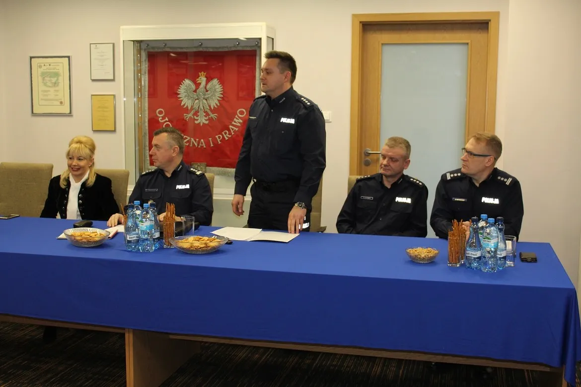Płoccy policjanci podczas konferencji w sali urzędu omawiają stan bezpieczeństwa w minionym roku, siedząc przy stole z wodą i przekąskami, z flagą w tle.