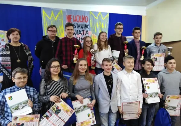 Grupa uczniów i opiekunów stoi na tle dekoracji, trzyma trofea i dyplomy po eliminacjach gminnych OTWP w Powiecie Płockim.