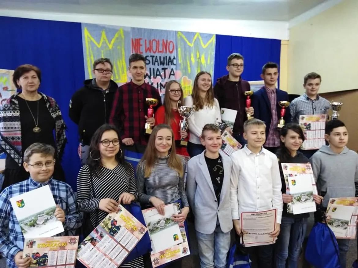 Grupa uczniów i opiekunów stoi na tle dekoracji, trzyma trofea i dyplomy po eliminacjach gminnych OTWP w Powiecie Płockim.