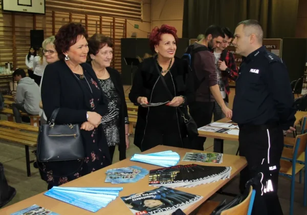 Uczestnicy IX Szkolnych Targ Edukacji i Pracy w Zespole Szkół im. St. Staszica w Gąbinie stoją przy stoisku z ulotkami i broszurami, rozmawiając z policjantem w mundurze.