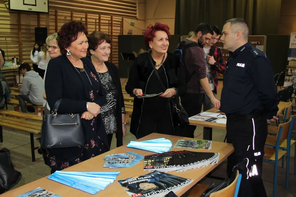 Uczestnicy IX Szkolnych Targ Edukacji i Pracy w Zespole Szkół im. St. Staszica w Gąbinie stoją przy stoisku z ulotkami i broszurami, rozmawiając z policjantem w mundurze.