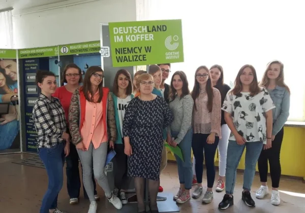 Grupa uczniów i nauczycielek Zespołu Szkół im. Leokadii Bergerowej w Płocku stoi razem w szkolnym holu, trzymając zielony plakat z napisem „DEUTSCHLAND IM KOFFER NIEMCY W WALIZCE”.