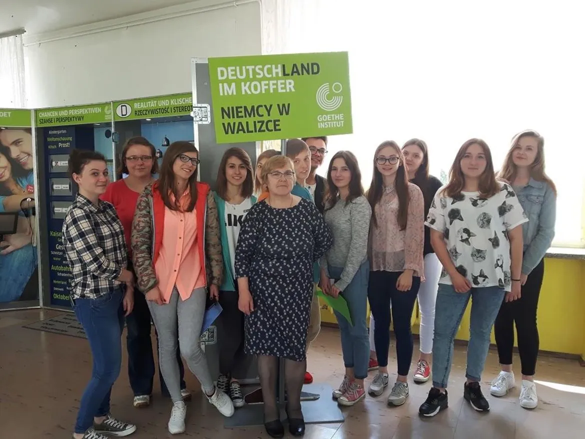 Grupa uczniów i nauczycielek Zespołu Szkół im. Leokadii Bergerowej w Płocku stoi razem w szkolnym holu, trzymając zielony plakat z napisem „DEUTSCHLAND IM KOFFER NIEMCY W WALIZCE”.