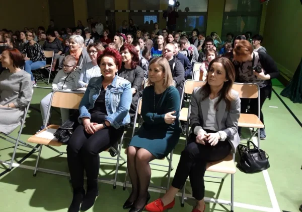 Widownia w hali sportowej podczas wydarzenia „NIKT NIE POZOSTAJE W TYLE” - w pierwszym rzędzie trzy kobiety siedzą na krzesłach, a za nimi zgromadzona jest duża grupa uczestników.