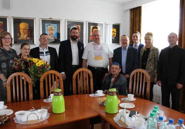 Grupa osób stoi w sali konferencyjnej, a w centrum widoczny mężczyzna z medalami olimpijskimi na szyi otrzymuje podziękowania dla mistrza olimpijskiego i jego trenera.