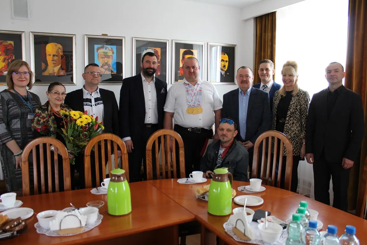 Grupa osób stoi w sali konferencyjnej, a w centrum widoczny mężczyzna z medalami olimpijskimi na szyi otrzymuje podziękowania dla mistrza olimpijskiego i jego trenera.