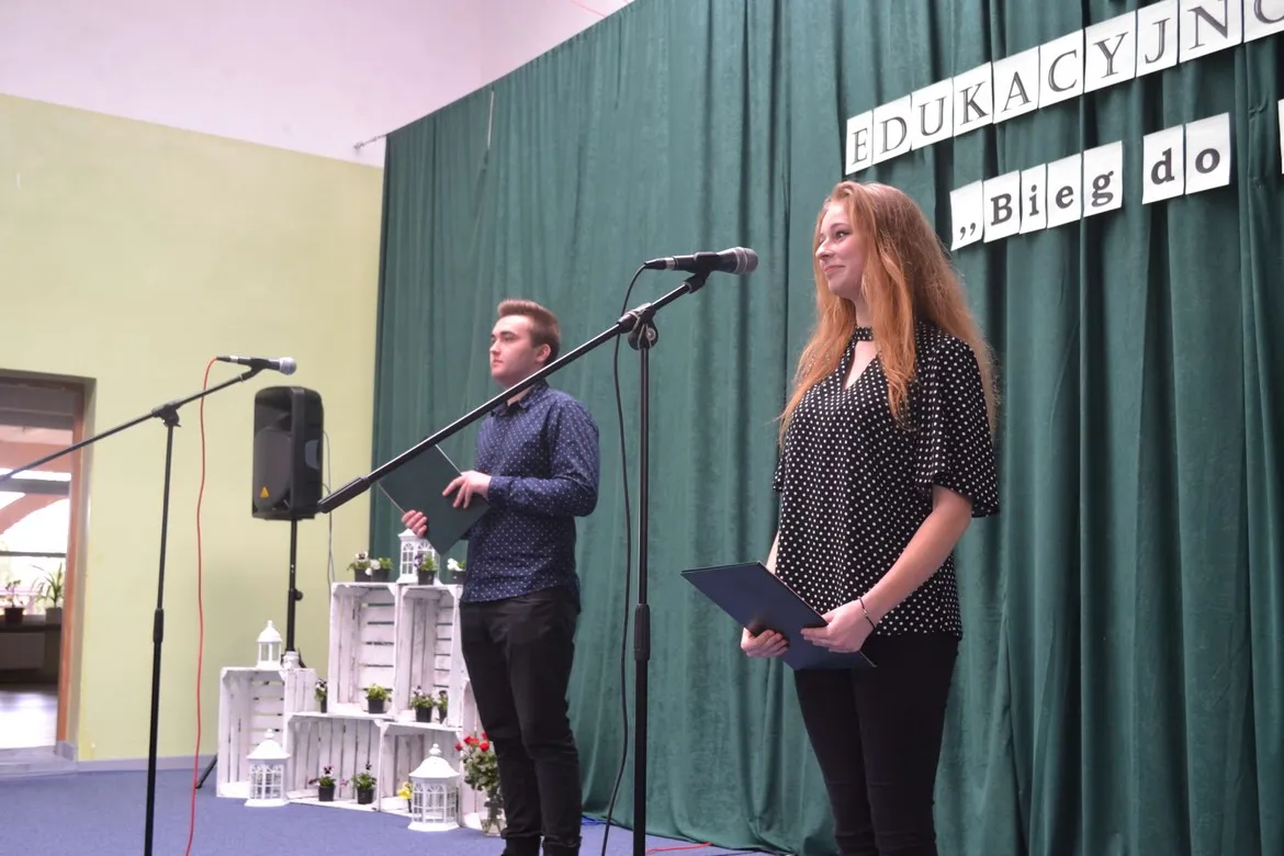 Dwóch młodych ludzi stoi na scenie podczas IV Targów Edukacyjno‑Zawodowych, przemawia do mikrofonów, a w tle widoczna jest zielona kurtyna i dekoracyjne białe skrzynie z lampionami.