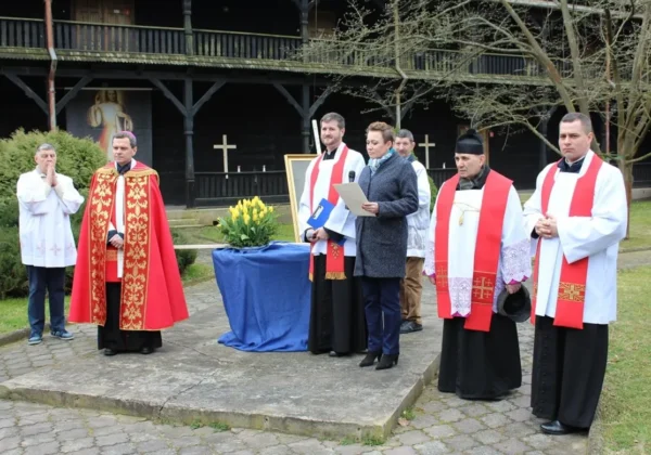 Grupa duchownych i uczestników Drogi Krzyżowej przed budynkiem DPS w Brwilnie, stojąca przy ołtarzu z żółtymi tulipanami; kapłani w białych albach z czerwonymi stułami, jedna osoba odczytuje tekst, w tle drewniana zabudowa i krzyże.