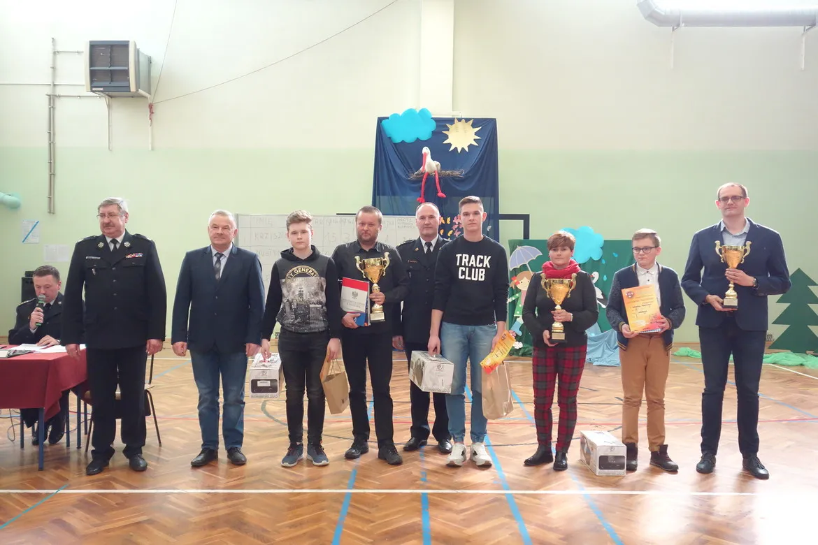Grupa uczestników i laureatów eliminacji powiatowych XLII edycji Ogólnopolskiego Turnieju Wiedzy Pożarniczej stoi na sali gimnastycznej, prezentując trofea, dyplomy i nagrody.