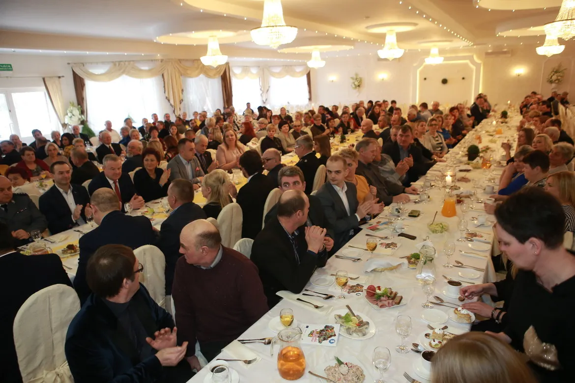 Duża hala bankietowa wypełniona gośćmi siedzącymi przy długich stołach zastawionych jedzeniem i napojami podczas spotkania Sołtysów Powiatu Płockiego – liderów rozwoju lokalnego.