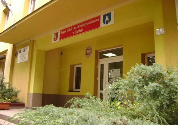 Wejście do Zespołu Szkół im. Stanisława Staszica w Gąbinie – żółty budynek z czerwoną tablicą z nazwą szkoły nad drzwiami i zielenią przed wejściem.