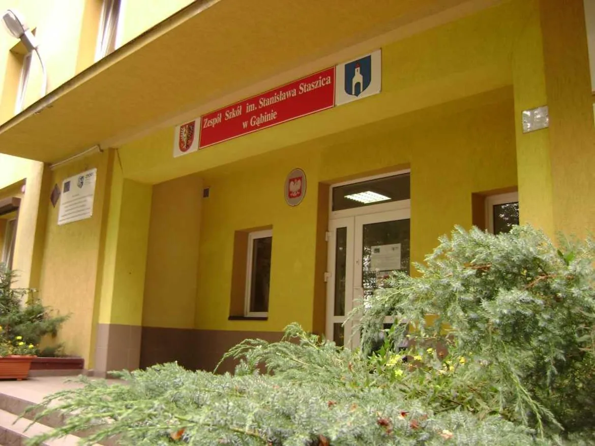Wejście do Zespołu Szkół im. Stanisława Staszica w Gąbinie – żółty budynek z czerwoną tablicą z nazwą szkoły nad drzwiami i zielenią przed wejściem.