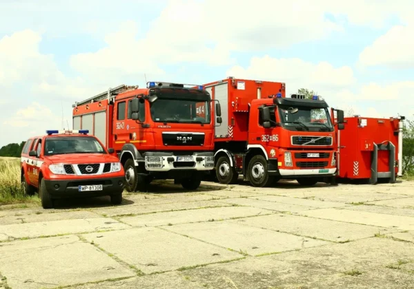 Trzy czerwone pojazdy straży pożarnej (radiowóz i dwa ciężkie samochody gaśnicze) stoją na betonowym placu obok mobilnego zestawu sprzętu ratowniczego.