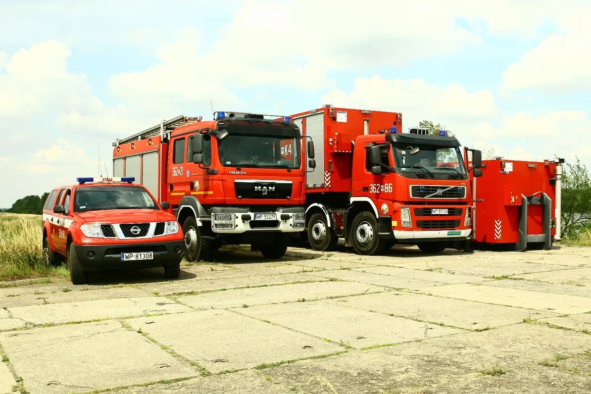 Trzy czerwone pojazdy straży pożarnej (radiowóz i dwa ciężkie samochody gaśnicze) stoją na betonowym placu obok mobilnego zestawu sprzętu ratowniczego.