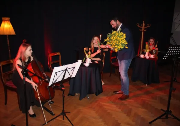 Podczas wieczorku muzycznego dla Pań i Panów kobieta gra na wiolonczeli, a mężczyzna wręcza bukiet żółtych kwiatów przy stoliku z czarnym obrusem.