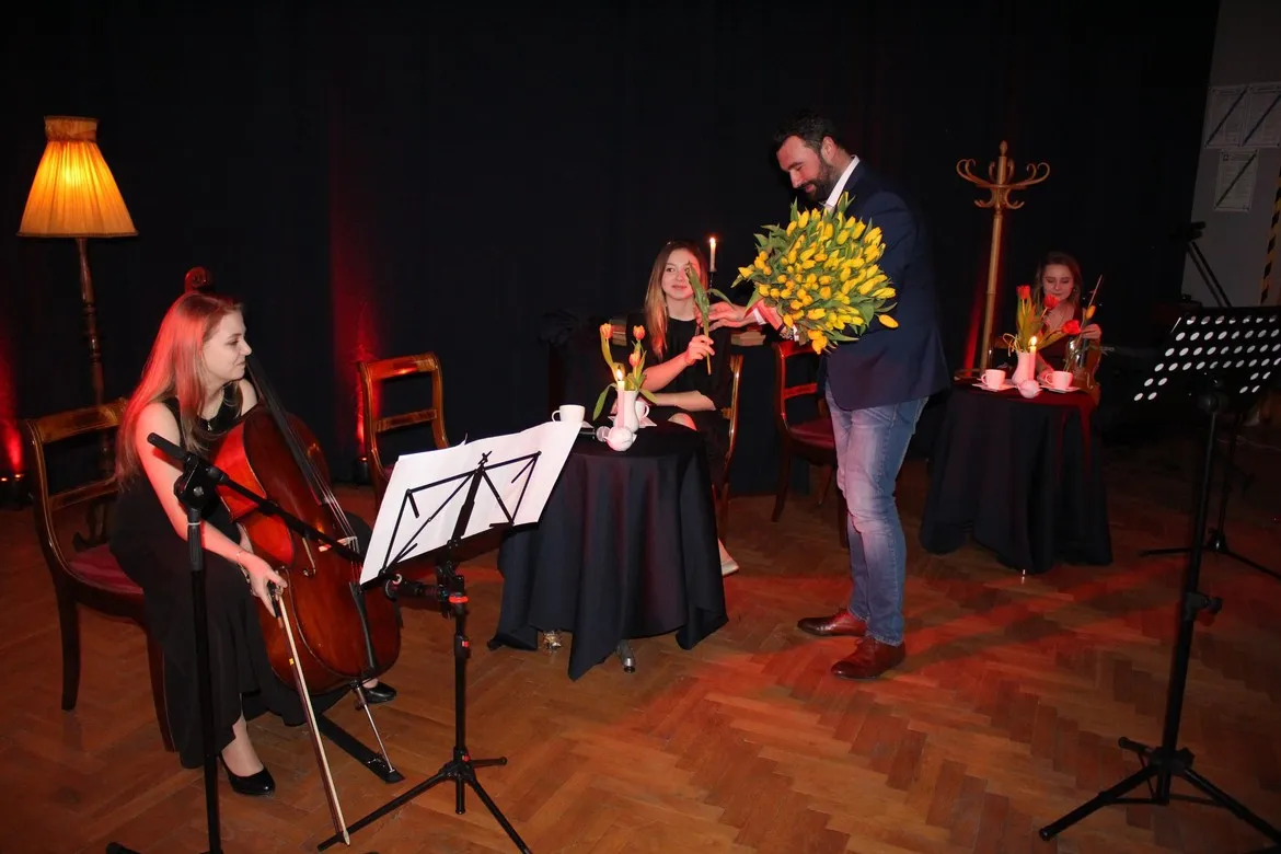 Podczas wieczorku muzycznego dla Pań i Panów kobieta gra na wiolonczeli, a mężczyzna wręcza bukiet żółtych kwiatów przy stoliku z czarnym obrusem.