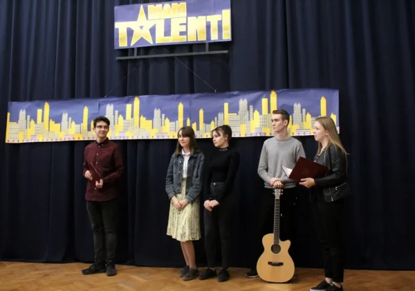 Pięć młodych ludzi stoi na scenie podczas szkolnego konkursu talentów „Mam Talent”; obok stoi gitara, a w tle widoczny jest baner z żółtymi sylwetkami miasta.