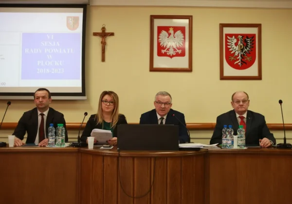 Czterech radnych siedzi przy stole podczas VI sesji Rady Powiatu w Płocku, z mikrofonami, dokumentami i laptopem, a w tle projekcja i oprawione na ścianie krzyż oraz dwa herby w ramkach.