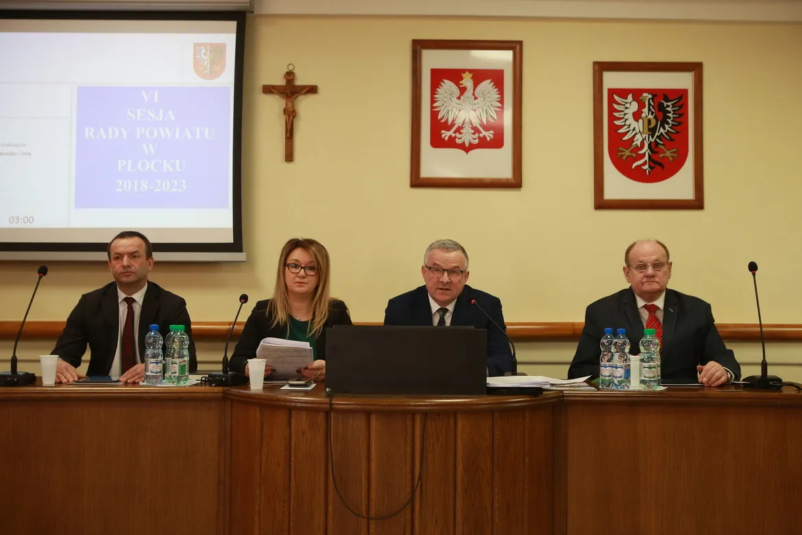 Czterech radnych siedzi przy stole podczas VI sesji Rady Powiatu w Płocku, z mikrofonami, dokumentami i laptopem, a w tle projekcja i oprawione na ścianie krzyż oraz dwa herby w ramkach.
