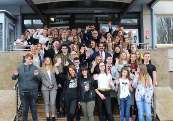 Duża grupa uśmiechniętych wolontariuszy WOŚP stoi na schodach przed budynkiem i macha do kamery w geście podziękowań.