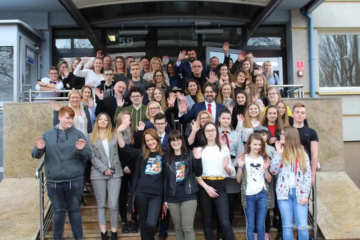 Duża grupa uśmiechniętych wolontariuszy WOŚP stoi na schodach przed budynkiem i macha do kamery w geście podziękowań.