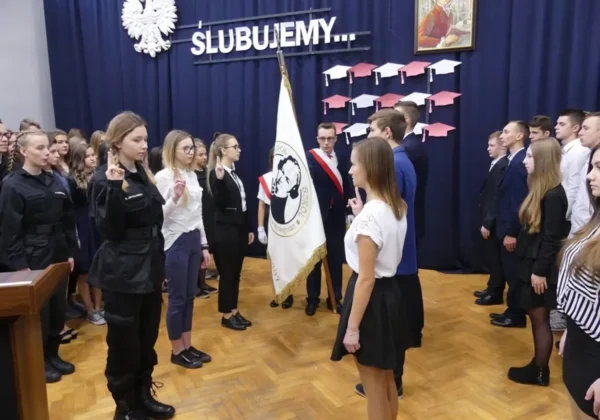 Uczniowie stoją w dwóch rzędach po obu stronach sali, składają przysięgę przed flagą szkoły, na tle dekoracji z napisem „ŚLUBUJEMY…”.