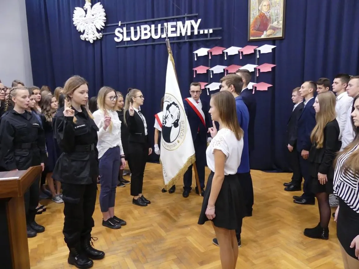 Uczniowie stoją w dwóch rzędach po obu stronach sali, składają przysięgę przed flagą szkoły, na tle dekoracji z napisem „ŚLUBUJEMY…”.