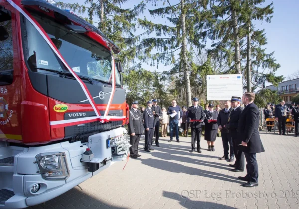 Uroczyste przekazanie nowego samochodu ratowniczo-gaśniczego Volvo podczas 96. rocznicy powstania OSP w Łęgu Probostwie; grupa strażaków w mundurach i gości stoi obok czerwonego pojazdu ozdobionego wstęgą.