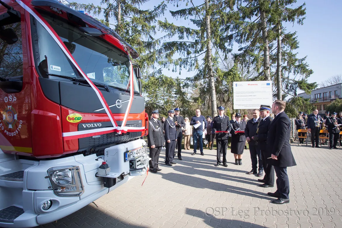 Uroczyste przekazanie nowego samochodu ratowniczo-gaśniczego Volvo podczas 96. rocznicy powstania OSP w Łęgu Probostwie; grupa strażaków w mundurach i gości stoi obok czerwonego pojazdu ozdobionego wstęgą.