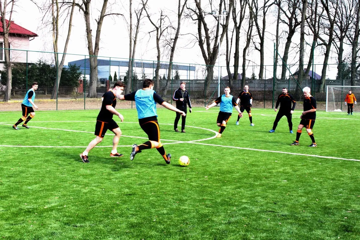 Grupa mężczyzn gra w piłkę nożną na nowym boisku z sztucznej trawy w Wyszogrodzie podczas jego otwarcia.