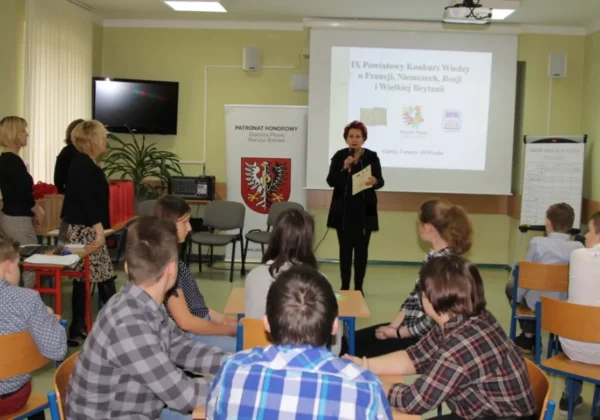 Prowadząca z mikrofonem przemawia podczas IX Powiatowego Konkursu Wiedzy o Francji, Niemczech, Rosji i Wielkiej Brytanii, a uczniowie siedzą przy stolikach w sali.