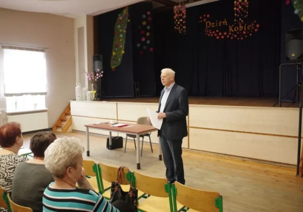 Mężczyzna w garniturze przemawia do zgromadzonej publiczności w sali, stojąc przy stole na scenie udekorowanej kwiatami i napisem Dzień Kobiet.