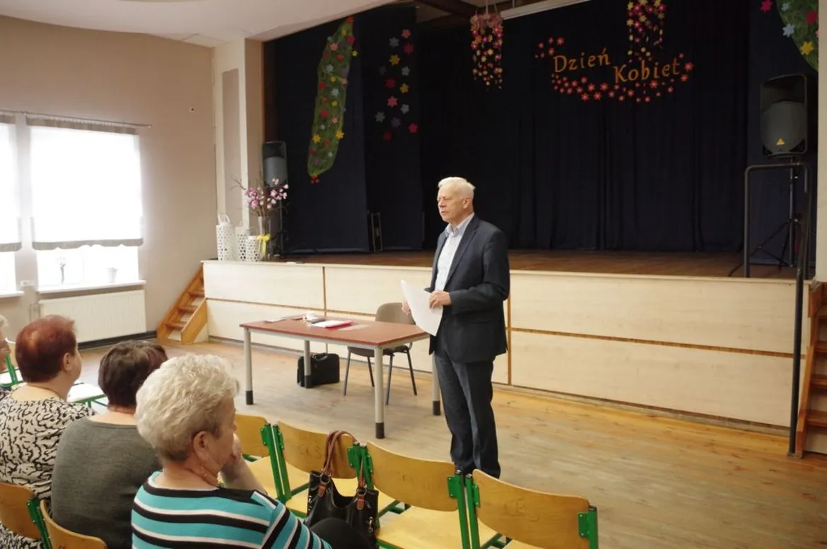 Mężczyzna w garniturze przemawia do zgromadzonej publiczności w sali, stojąc przy stole na scenie udekorowanej kwiatami i napisem Dzień Kobiet.