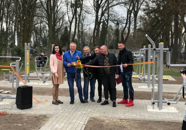 Grupa osób podczas otwarcia siłowni plenerowej wybudowanej przez Stowarzyszenie z Wyszogrodu, przecinająca pomarańczową wstęgę na tle urządzeń do ćwiczeń w parku.