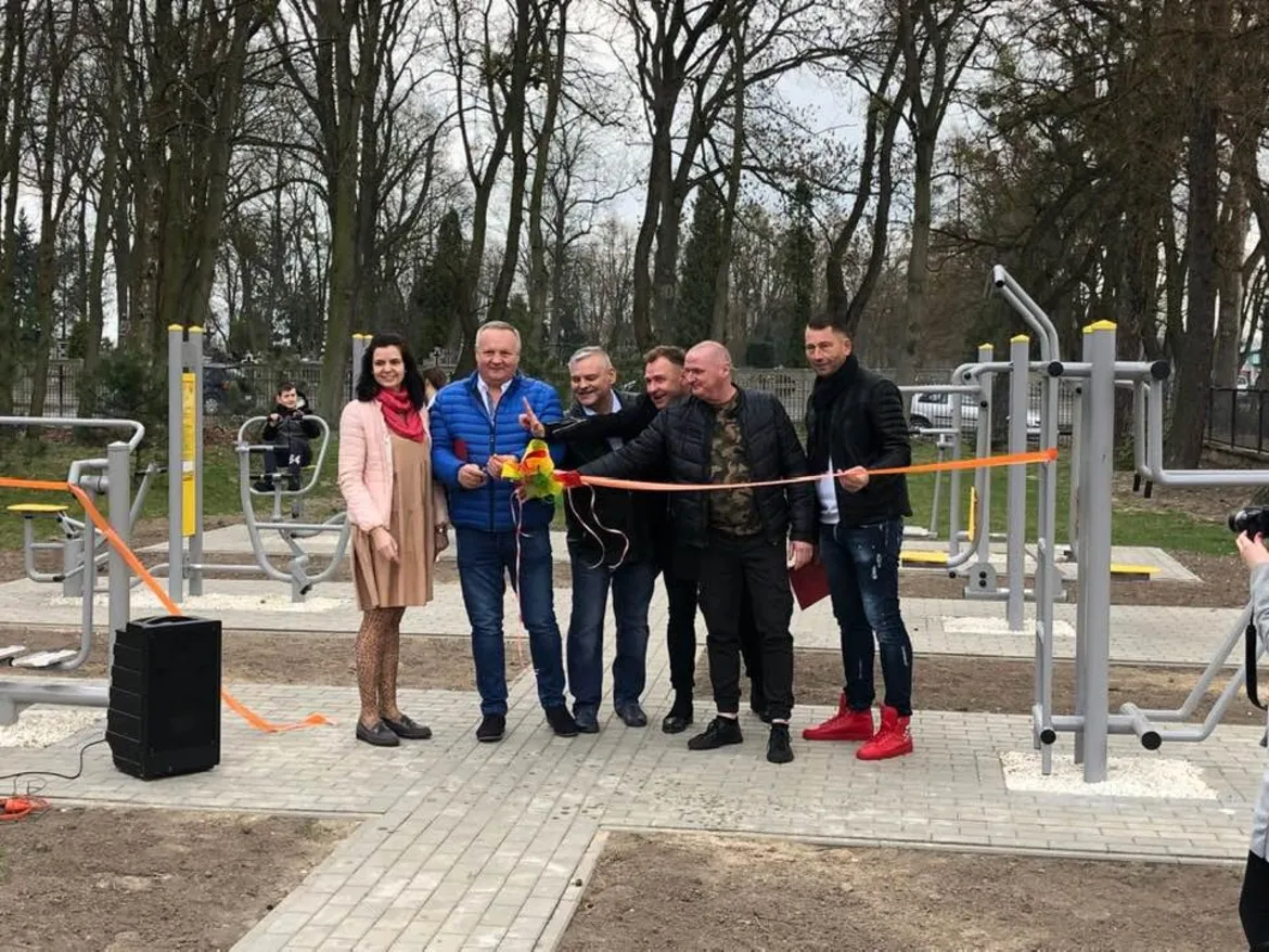 Grupa osób podczas otwarcia siłowni plenerowej wybudowanej przez Stowarzyszenie z Wyszogrodu, przecinająca pomarańczową wstęgę na tle urządzeń do ćwiczeń w parku.