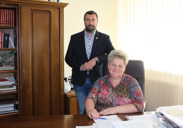 Mężczyzna w ciemnym garniturze stoi za siedzącą przy biurku kobietą w biurze powiatu, oboje uśmiechnięci.