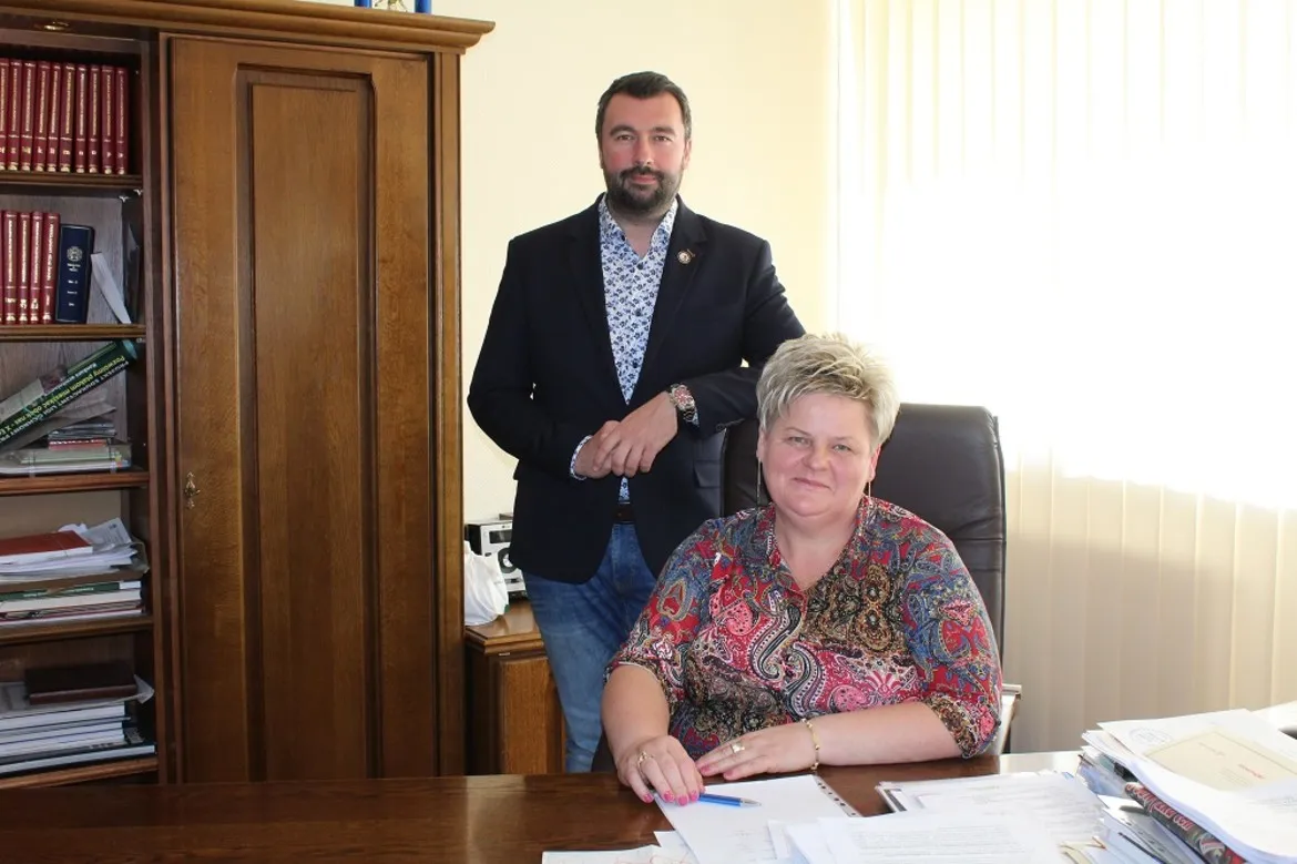 Mężczyzna w ciemnym garniturze stoi za siedzącą przy biurku kobietą w biurze powiatu, oboje uśmiechnięci.